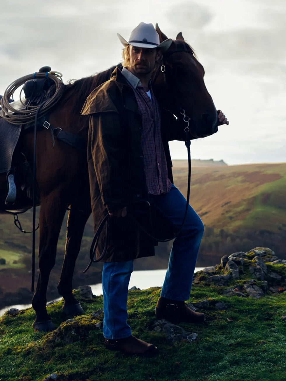 A man in a western cowboy outfit with a horse and a background of rolling hills.