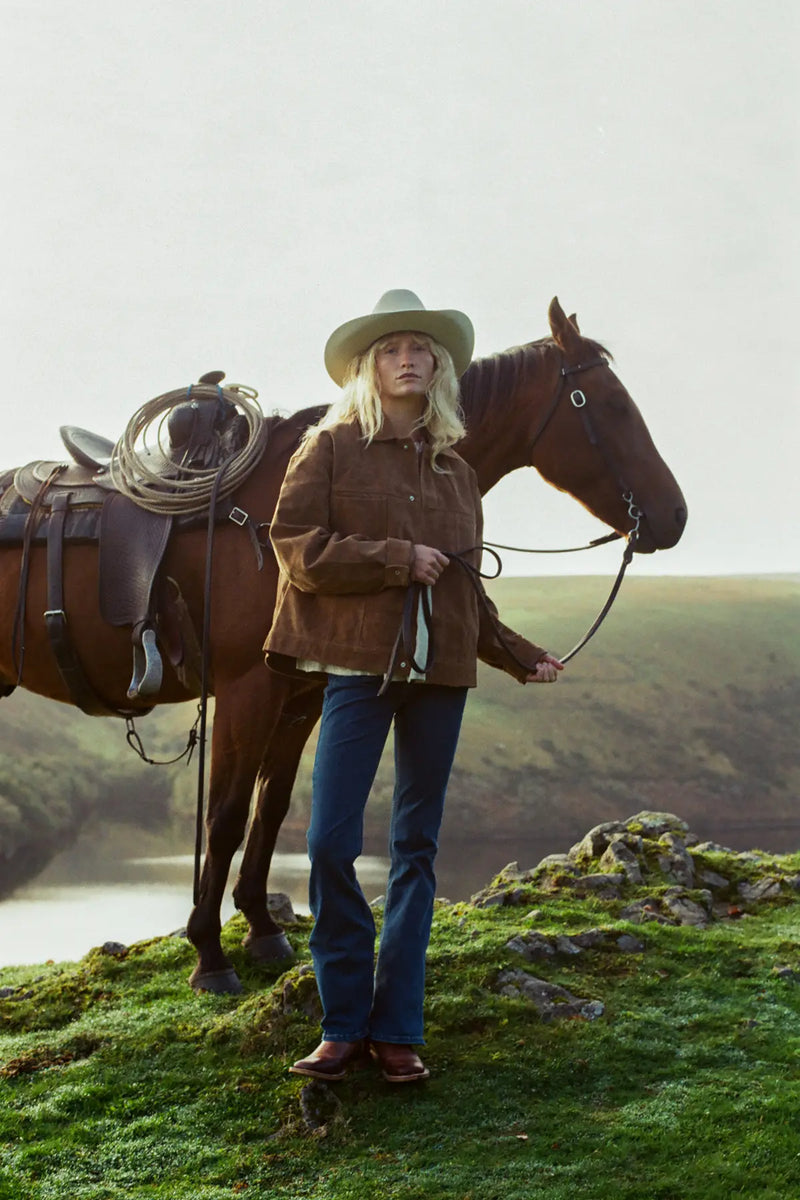Jenna in a cowboy hat and leather jacket standing next to a horse on a grassy hill with a scenic background.