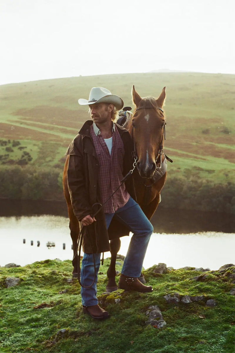 Matthew in a cowboy hat standing next to a horse by a lake with a scenic background