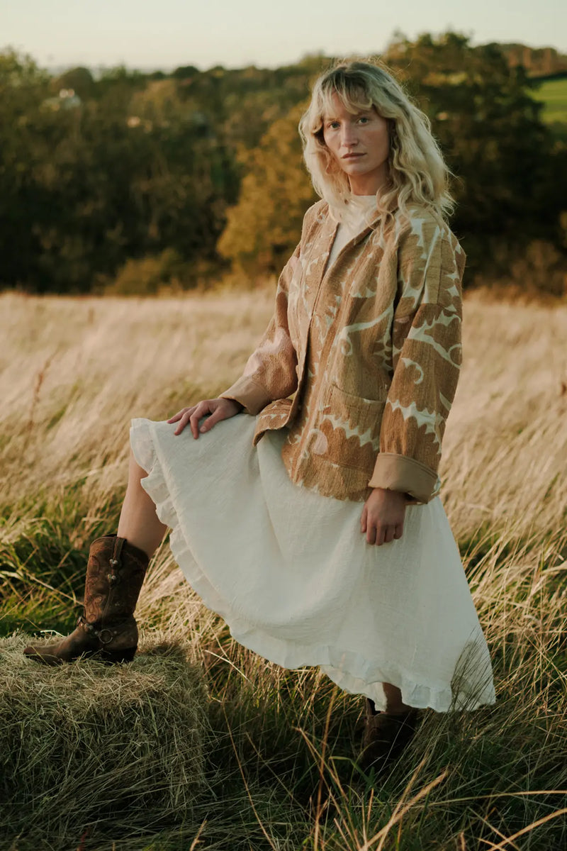 Jenna in a patterned jacket, white dress and Durango boots standing in a field with foot on a haybale and trees in the background