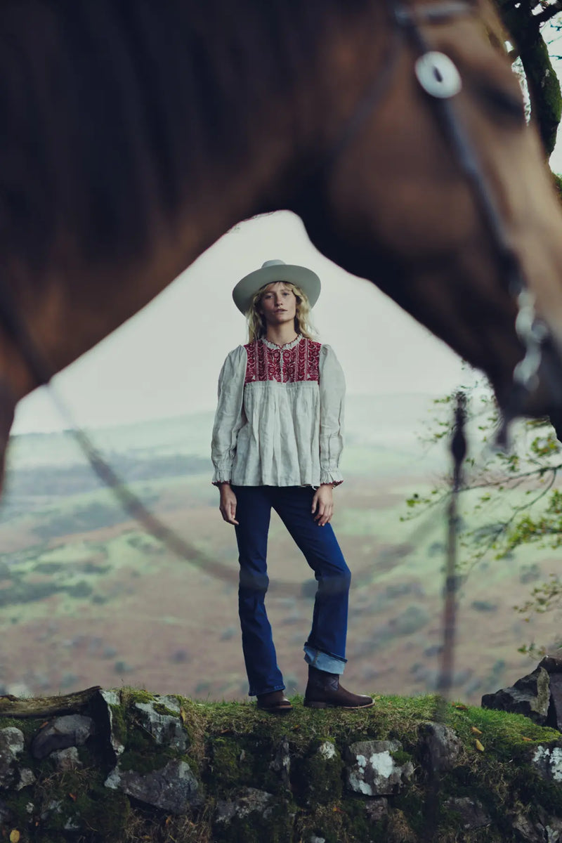 Jenna in a patterned blouse, jeans and Durango boots standing in front of a horse with a scenic background.