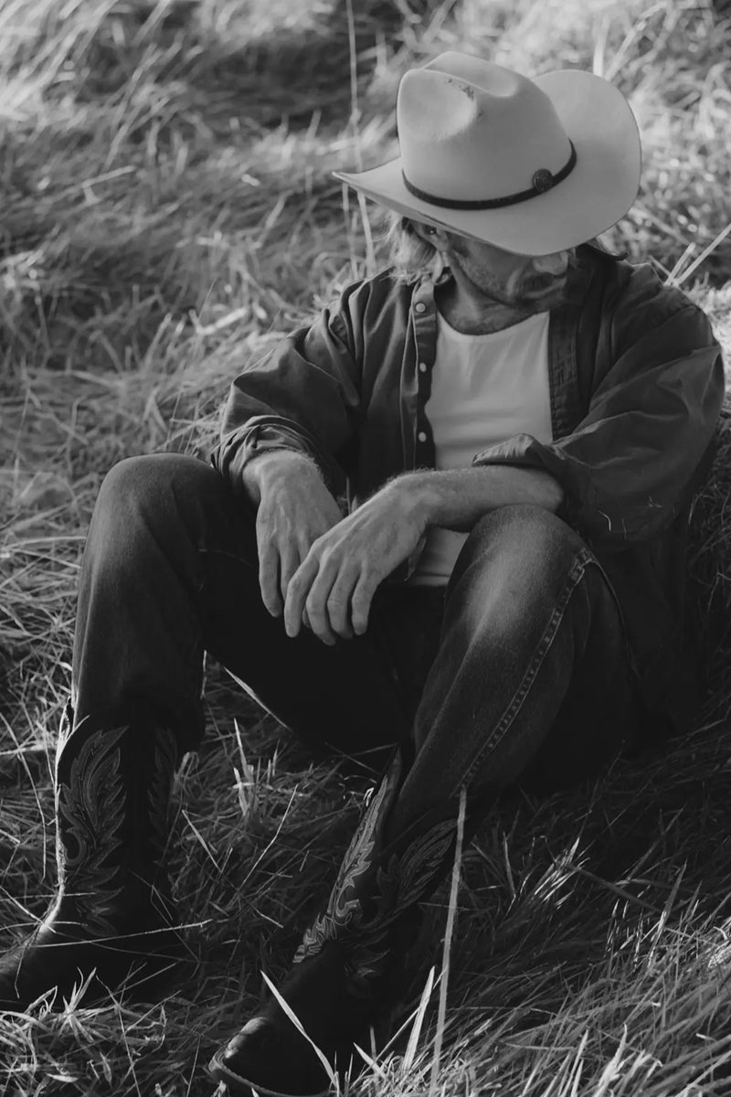 Matthew sitting in a field wearing a cowboy hat and Durango boots.