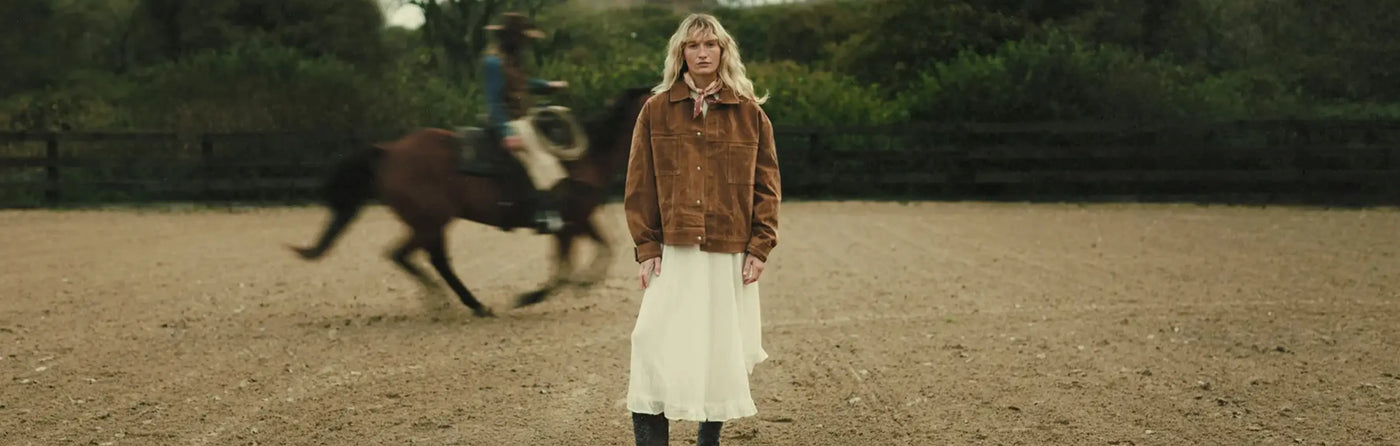 Woman in a white dress wearing a pair of Durango western boots standing in an equestrian arena with a blurred horse running in the background.