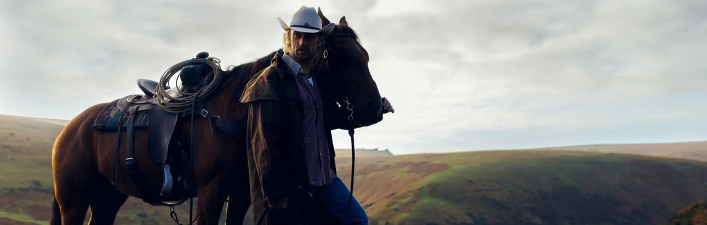 A man in a western cowboy outfit with a horse and a background of rolling hills.