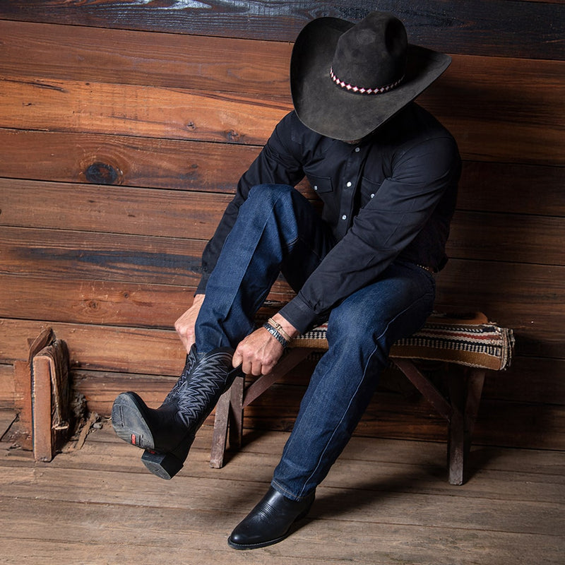 Man sat on a bench, wearing a black cowboy hat and putting on a pair of black Durango Rebel Frontier Western Boots